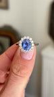 Antique-inspired intense cornflower blue sapphire and diamond cluster engagement ring showcasing an oval cornflower blue sapphire, diamond halo and elegant gallery setting, held by hand.