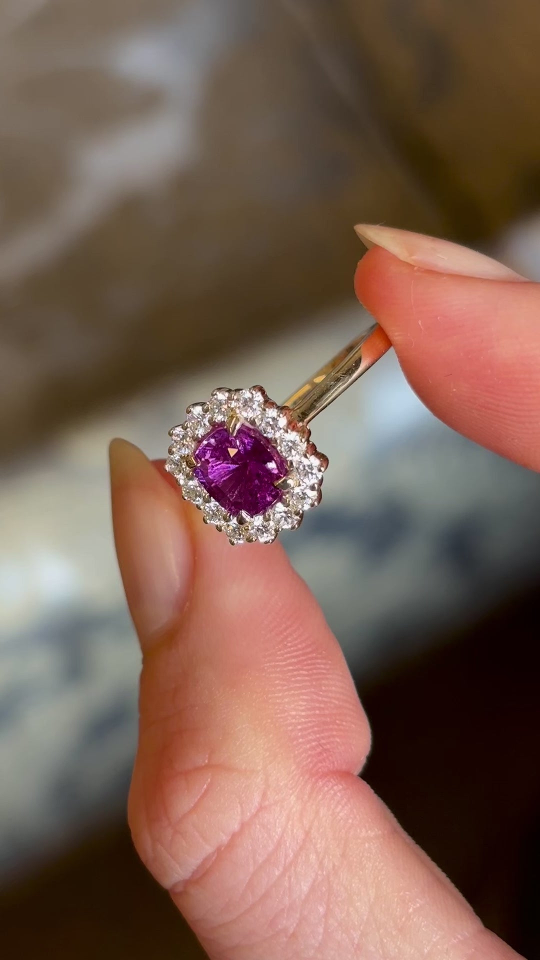 Square-cut pink sapphire and diamond cluster engagement ring held in fingers and moved slowly from side to side 