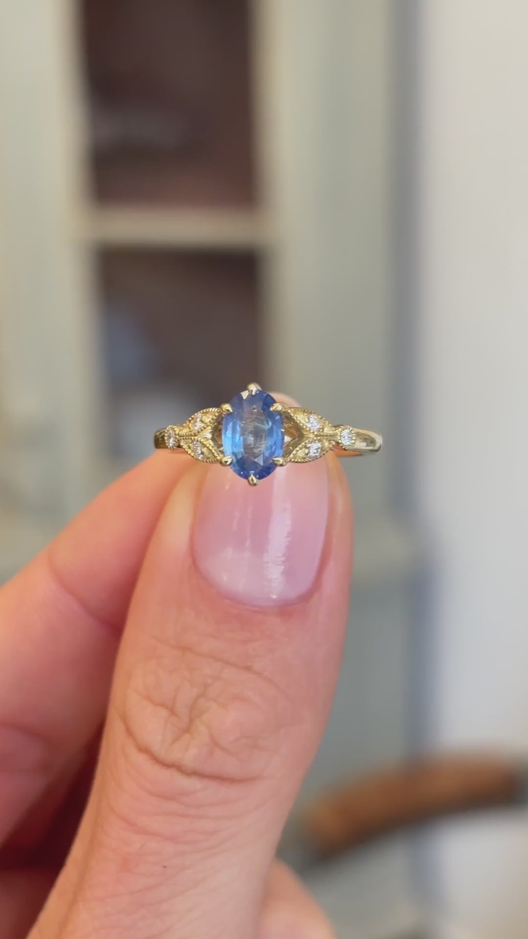 Blue sapphire and diamond engagement ring showcasing an oval blue sapphire centre stone with diamond-set trefoil shoulders in the form of laurel leaves, held by hand to highlight the setting and craftsmanship.
