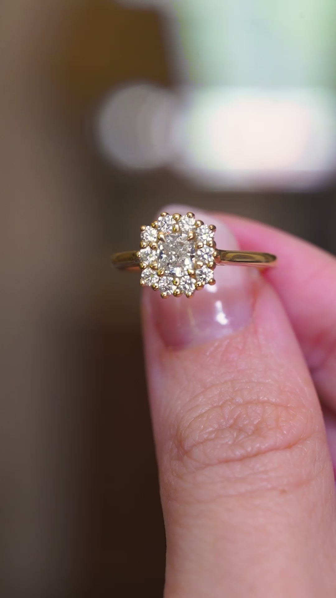A close-up of a vintage-inspired cushion-cut diamond halo engagement ring in yellow gold shown in motion between fingers.