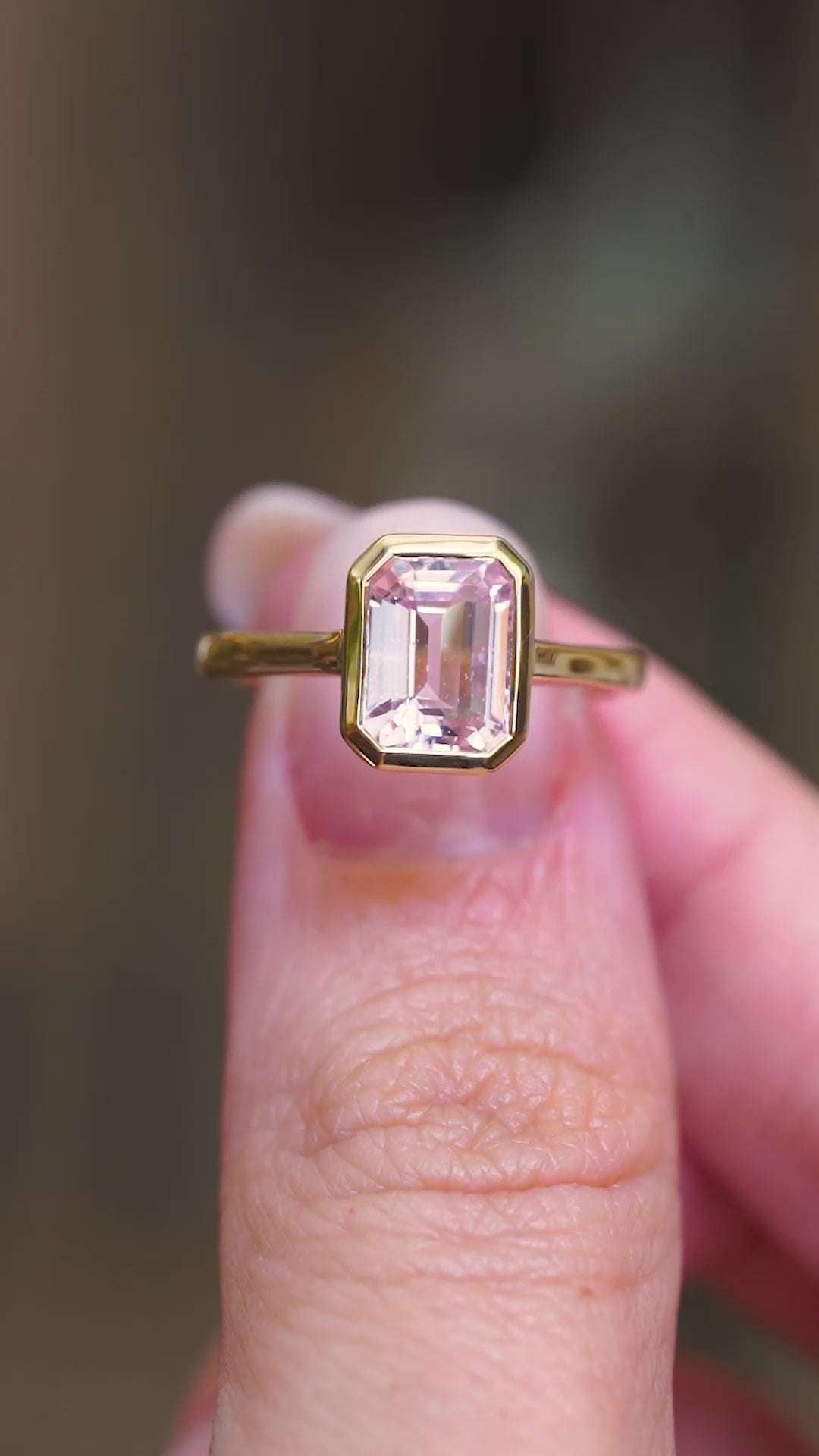 A vintage-inspired, soft petal-pink sapphire solitaire engagement ring in 14ct yellow gold, held between fingers showing the ring details. 