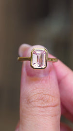 A vintage-inspired, soft petal-pink sapphire solitaire engagement ring in 14ct yellow gold, held between fingers showing the ring details. 