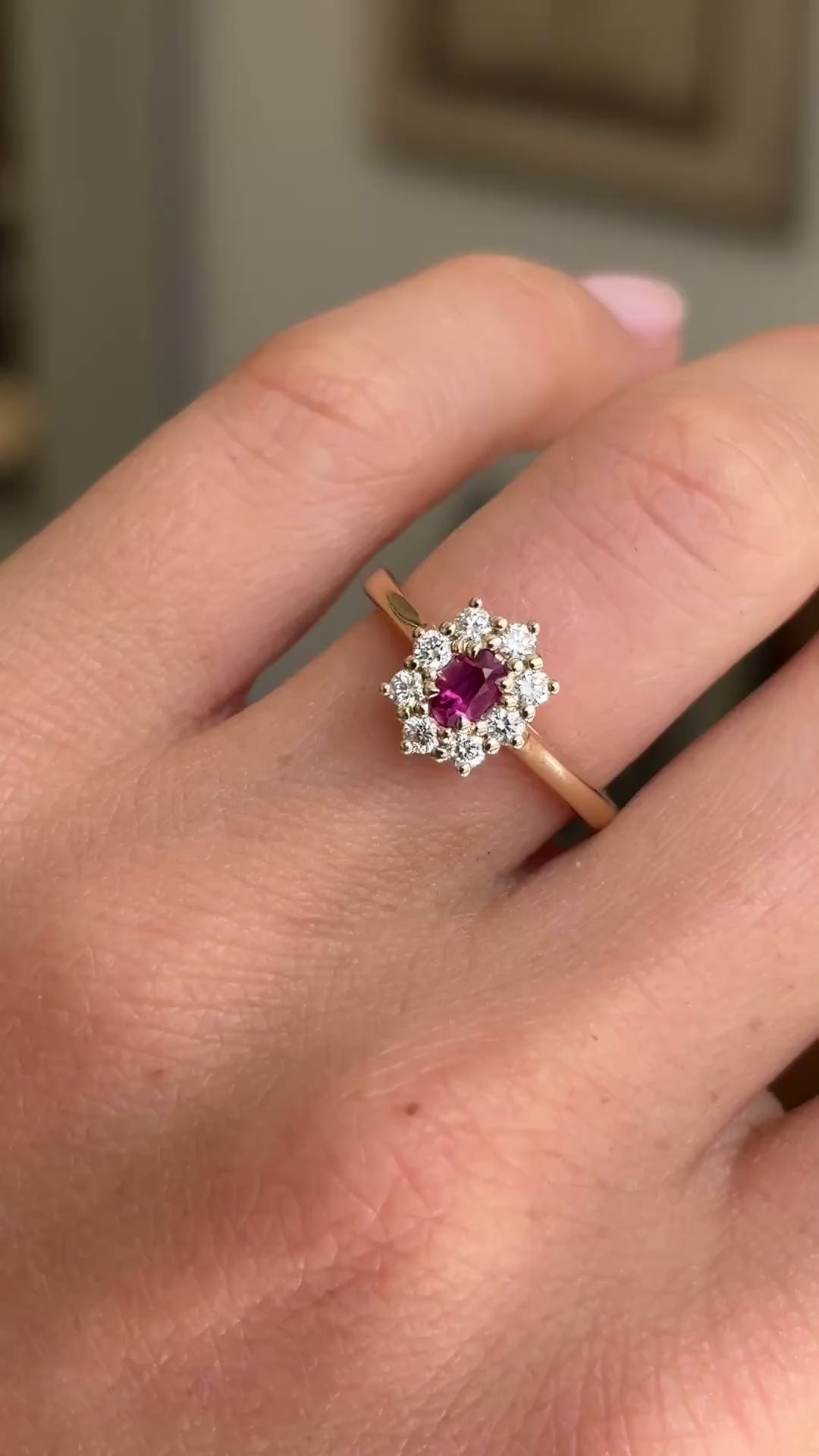 Detailed video of an oval-cut ruby and diamond cluster ring in yellow gold, held between fingers to showcase the vibrant ruby centre stone, diamond halo and elegant antique-inspired design against a warm interior setting.