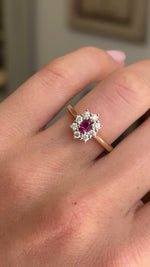 Detailed video of an oval-cut ruby and diamond cluster ring in yellow gold, held between fingers to showcase the vibrant ruby centre stone, diamond halo and elegant antique-inspired design against a warm interior setting.