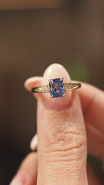 A Vintage-inspired, Blue sapphire and diamond flanked yellow gold solitaire engagement ring heldheld between two fingers and shown in detail as the ring is slowly rotated to show off different angles.