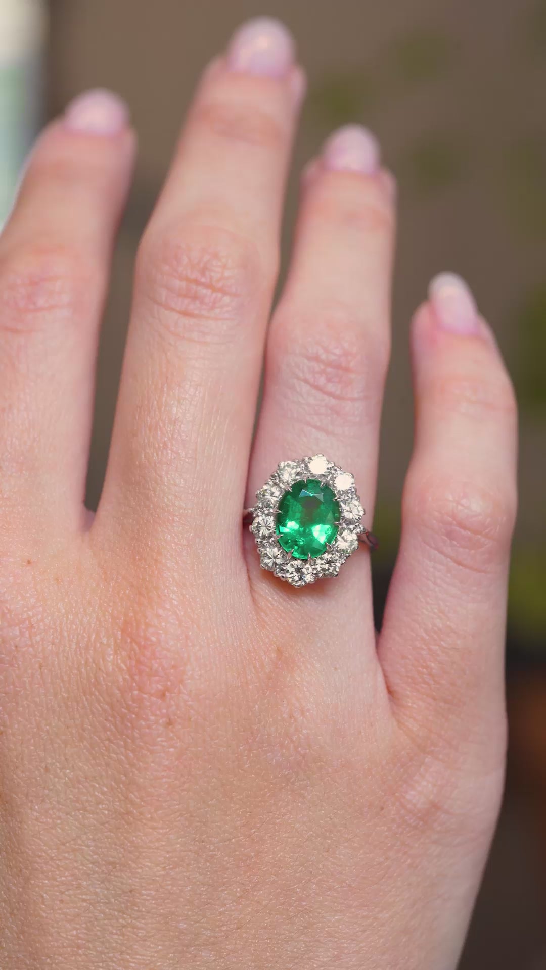 A video of a vintage 1950s oval-cut emerald and diamond cluster engagement ring in white gold worn on a hand.