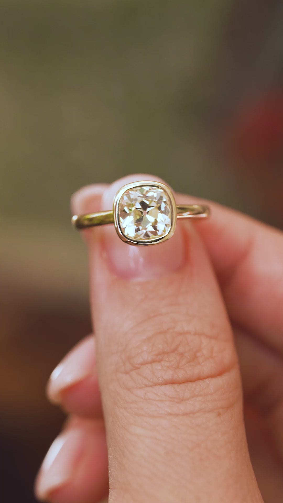 Video close-up of an old-mine cut diamond ring balanced on a fingertip, displaying the cushion stone and polished gold surround.