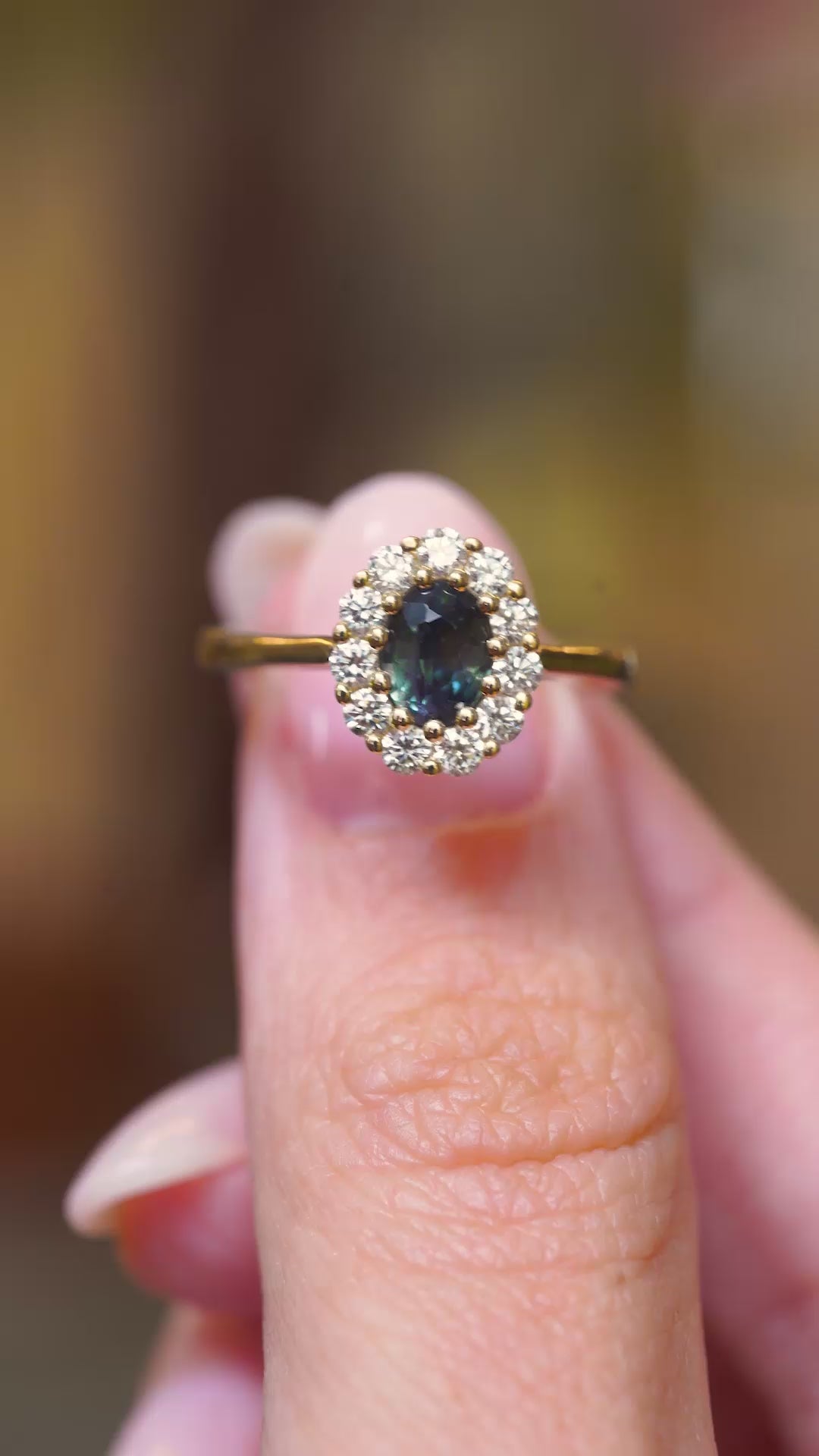 A close-up video of an oval deep green sapphire framed by a sparkling diamond cluster in an antique-inspired yellow gold engagement ring held between fingers.