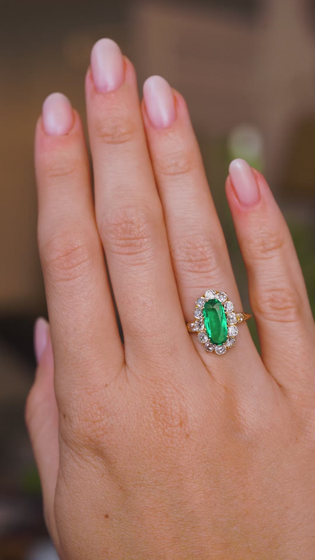 A video of a hand wearing an Antique, Belle Époque emerald and diamond cluster engagement ring.  Rotated slowly to show the profile of the ring once worn and the yellow gold band.  