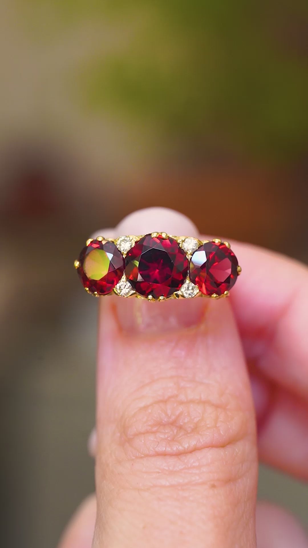 A vintage three-stone garnet with a deep red hue set in carved yellow gold showing in video between fingers.