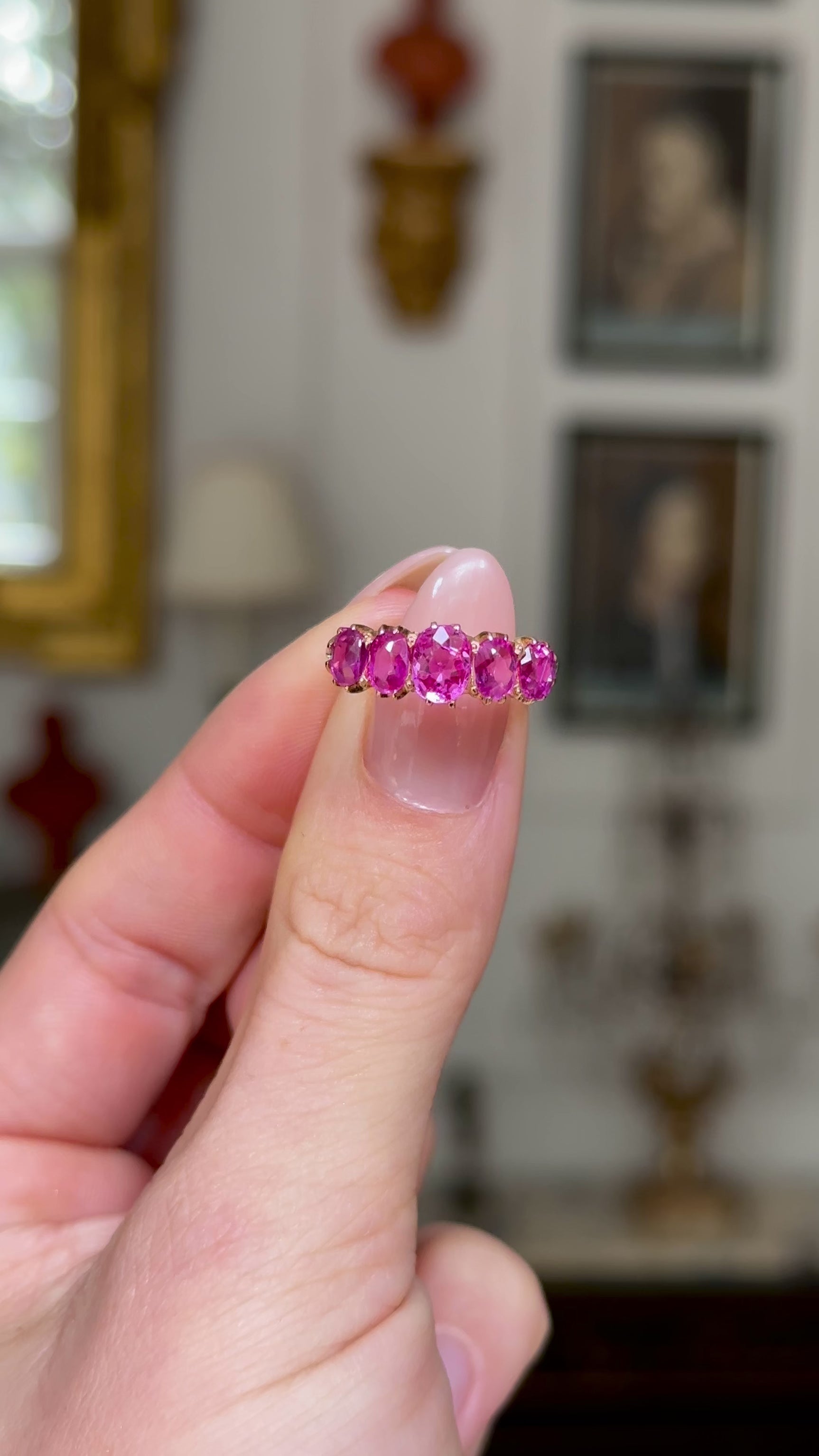 Five stone pink sapphire and yellow gold ring, held in fingers and rotated to provide full perspective. shot on antiquated bokeh background.