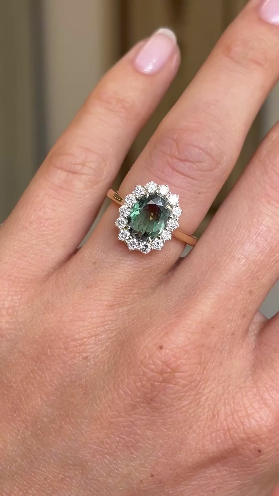 A video of a hand wearing a yellow gold engagement ring centred with a unique colour-change alexandrite with a halo of round brilliant diamonds. 