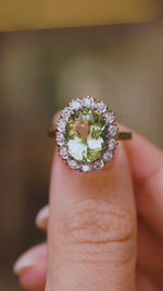 A close up video of a Vintage, 1950s green beryl and diamond cluster engagement ring held between two fingers and rotated slowly to display the front and side profiles of the ring and yellow gold band. 