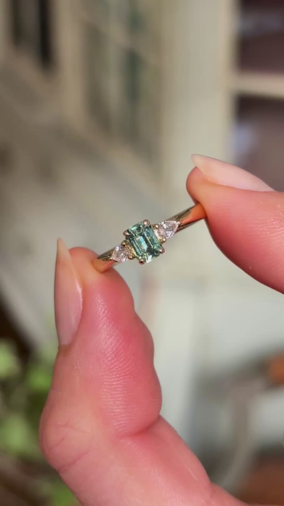 Video of a green sapphire and diamond three-stone engagement ring held between fingers in natural light, highlighting gemstone colour and brilliance.