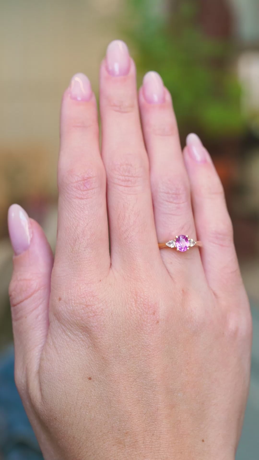 Pink oval sapphire and diamond three-stone engagement ring worn on hand with subtle motion.
