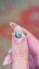 Teal sapphire and diamond cluster tre-foil engagement ring held between fingers showing ring details.