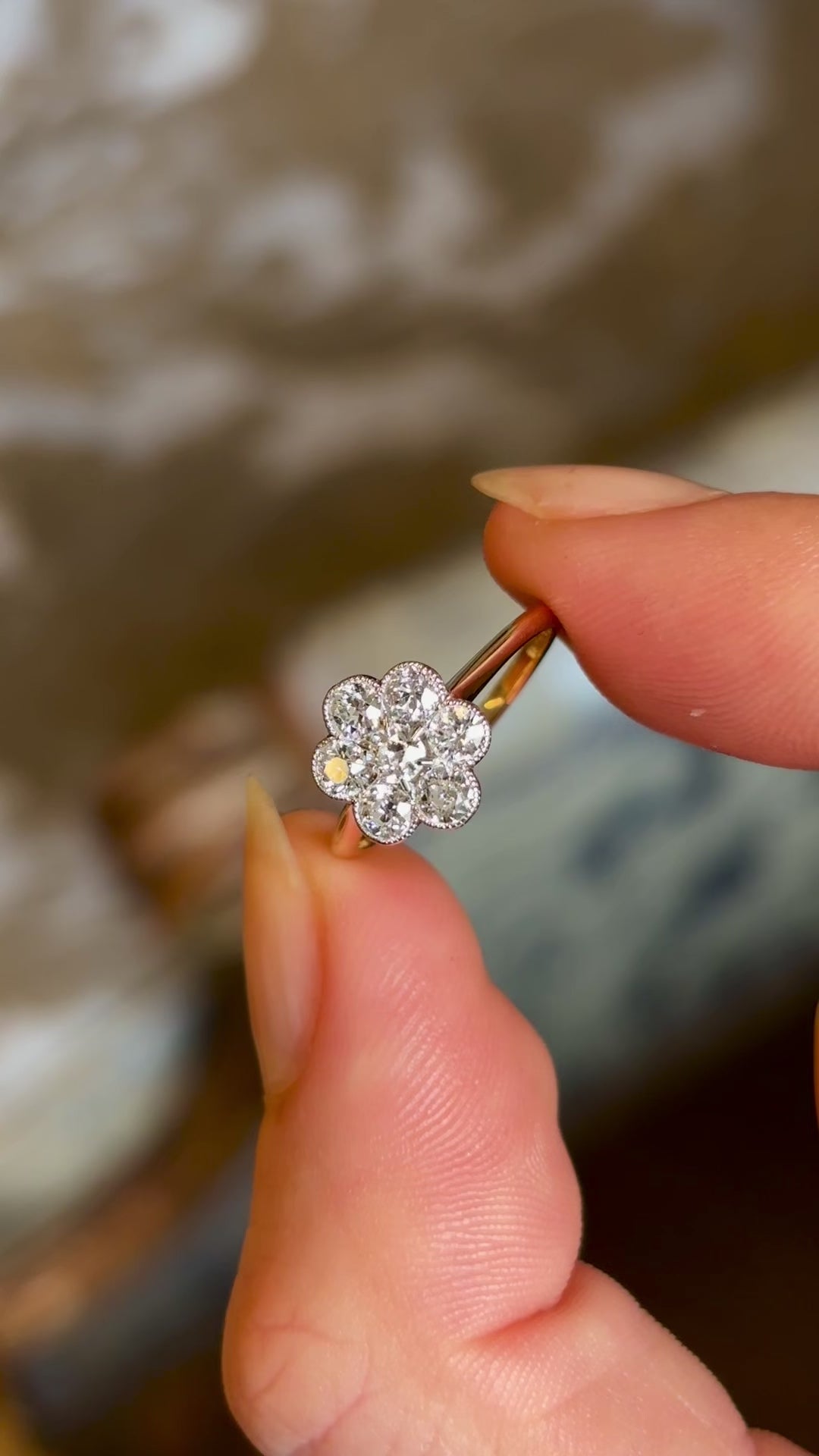 Two fingers holding an Edwardian daisy diamond cluster ring with an 18ct yellow gold band with a blurred background. 