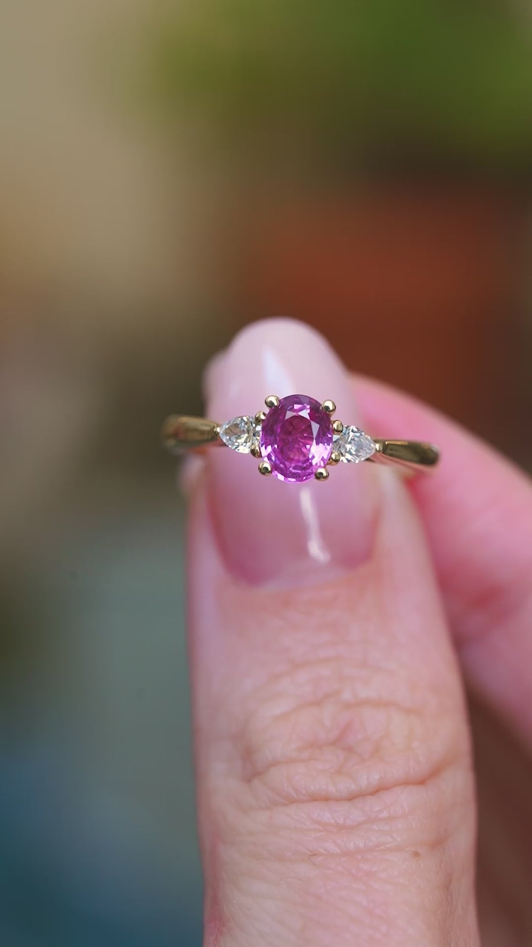 Pink oval sapphire and diamond three-stone engagement ring held between fingers showing ring details.