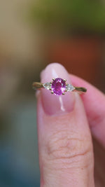 Pink oval sapphire and diamond three-stone engagement ring held between fingers showing ring details.