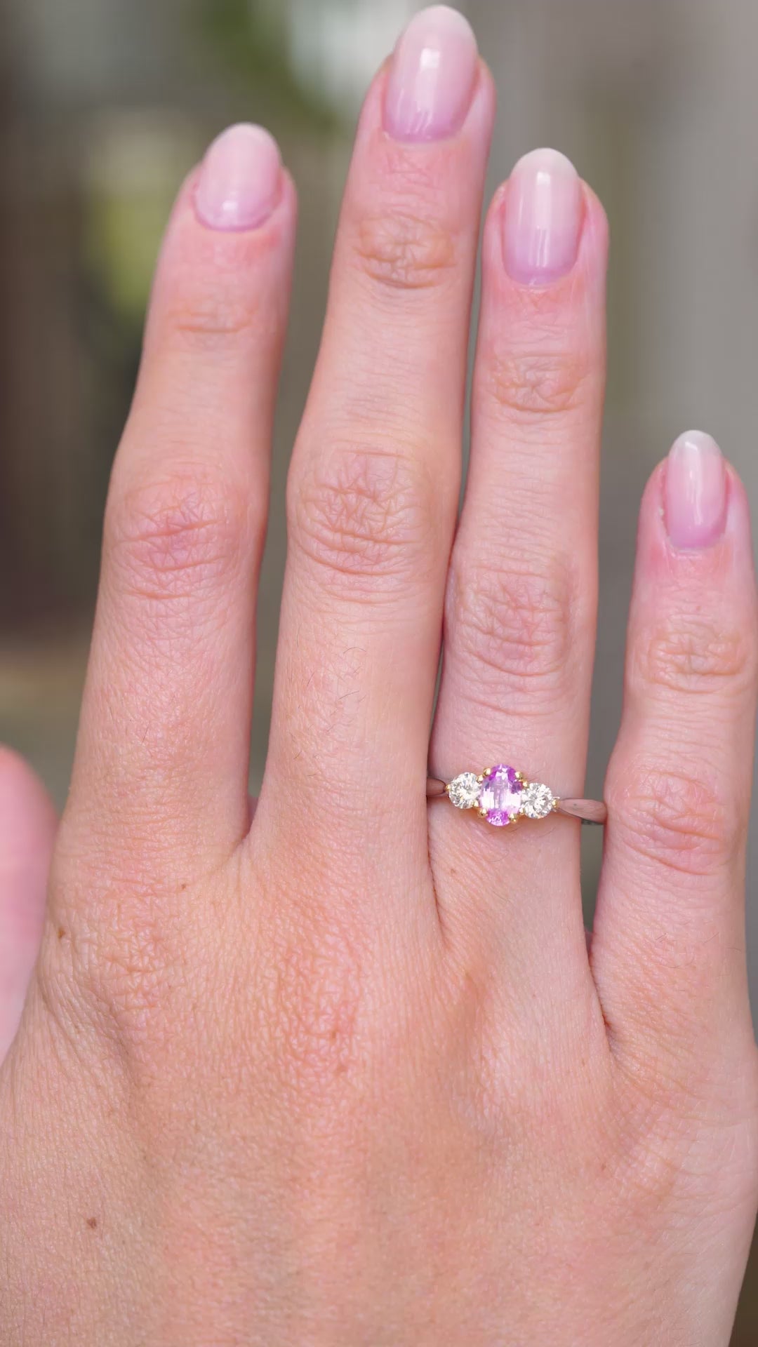 A video of a 1990s Vintage platinum three stone engagement ring being worn on the hand against a blurred background, showcasing the pink sapphire and diamonds. 