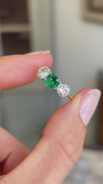 Antique Edwardian emerald and diamond three-stone engagement ring featuring an emerald centre stone flanked by two diamonds, held by hand for detailed viewing.