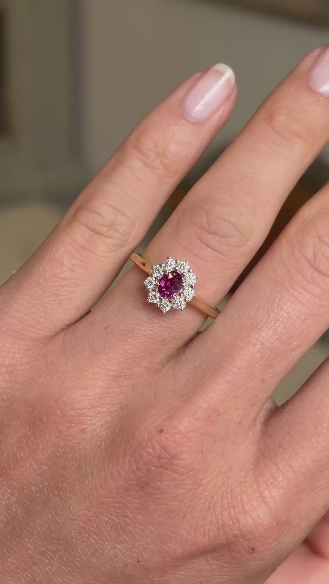 A hand wearing an antique-inspired cluster engagement ring with a round pinkish-red central ruby surrounded by a halo of diamonds, on a gold band shown in video.
