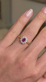 A hand wearing an antique-inspired cluster engagement ring with a round pinkish-red central ruby surrounded by a halo of diamonds, on a gold band shown in video.