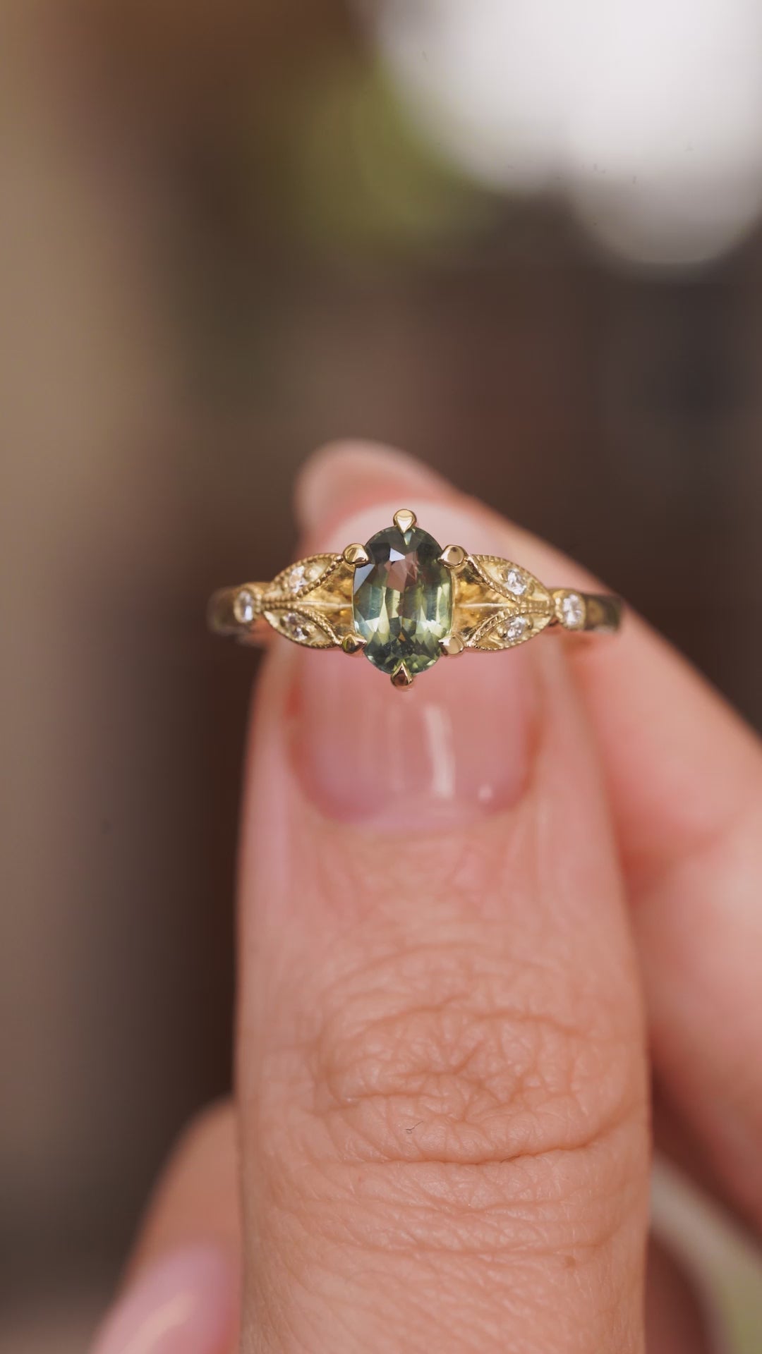 A video of a Vintage-inspired, Green sapphire and diamond engagement ring held in between the fingers