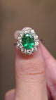 A video of an oval-cut emerald surrounded by a diamond cluster in a vintage 1950s white gold engagement ring held between fingers.
