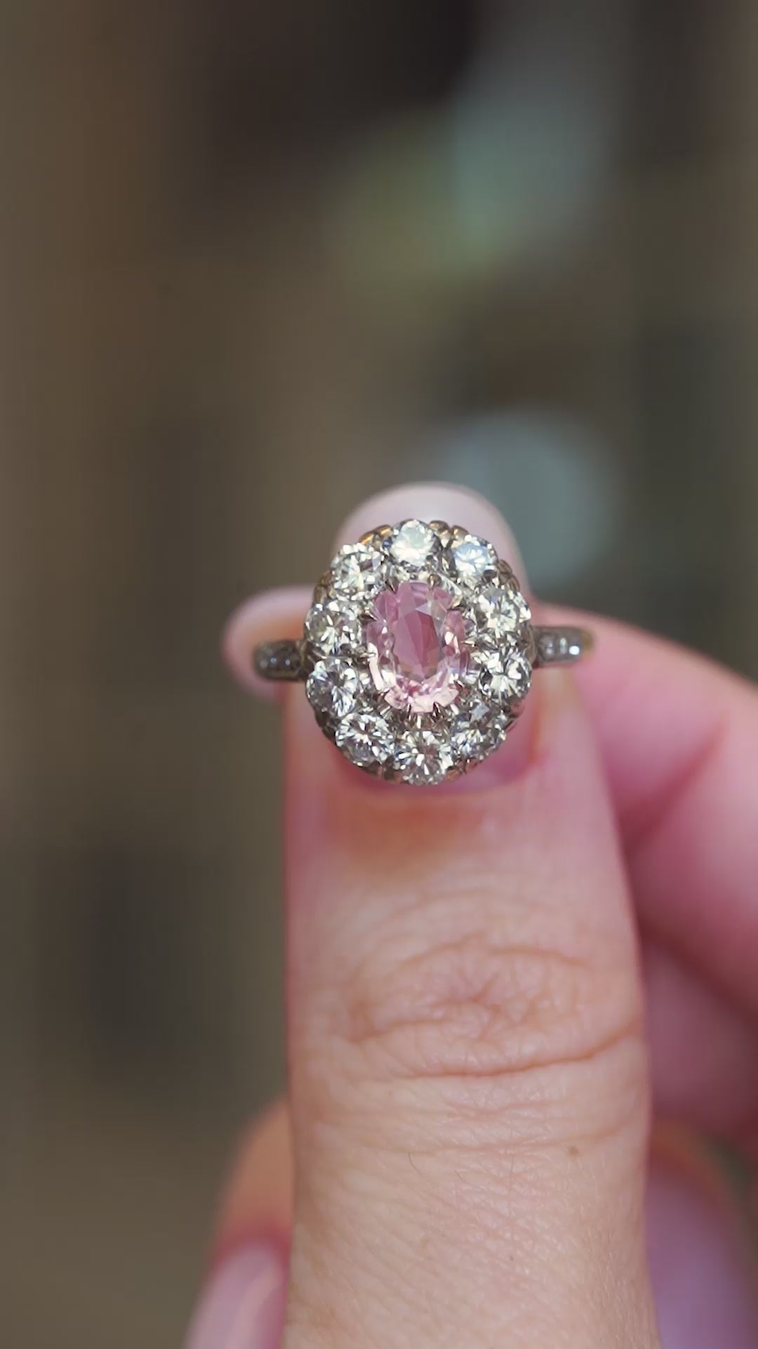 An oval orange-pink sapphire framed by a halo cluster old-cut diamonds shown between fingers.
