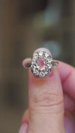 An oval orange-pink sapphire framed by a halo cluster old-cut diamonds shown between fingers.