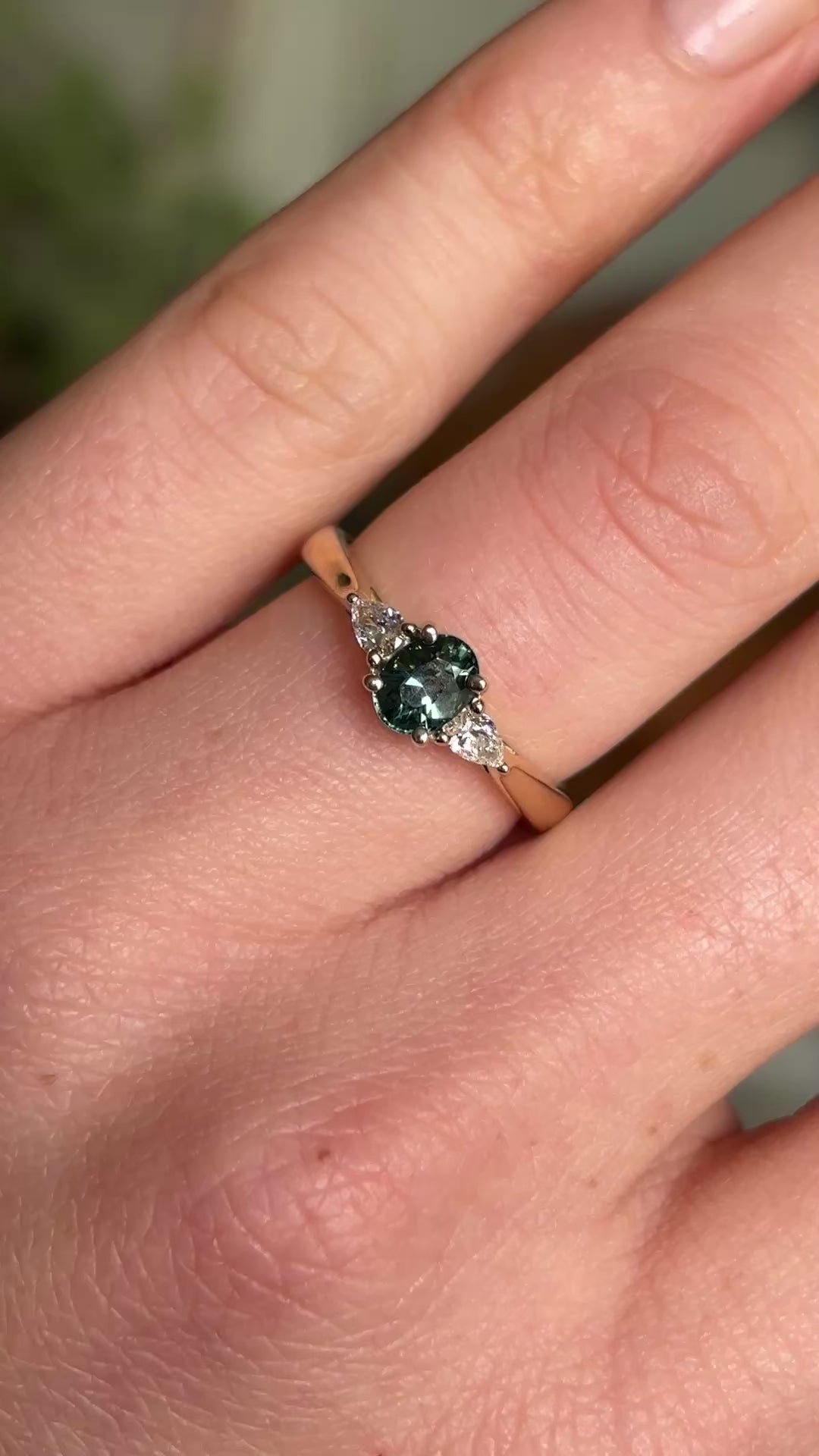 Oval-cut teal sapphire and diamond three-stone engagement ring worn on the hand, emphasising the sapphire’s colour and diamond accents.