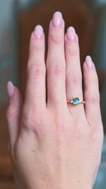 Teal sapphire and diamond three-stone engagement ring worn on hand with gold band with subtle motion.