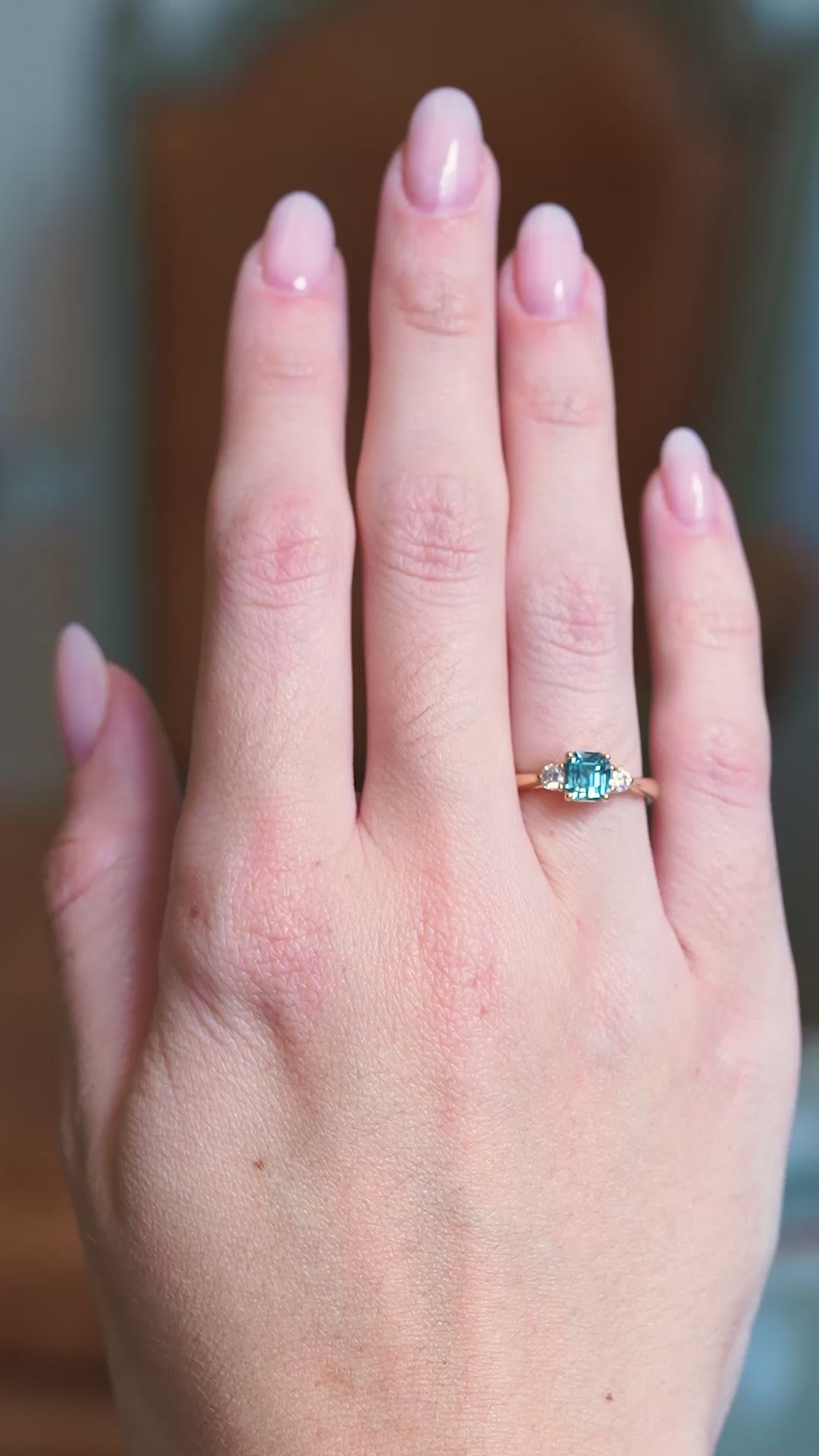 Teal sapphire and diamond three-stone engagement ring worn on hand with gold band with subtle motion.