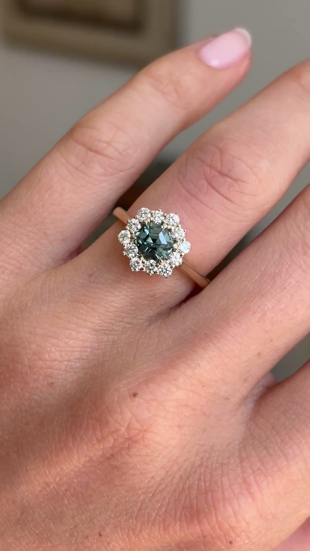 Teal hexagon-cut sapphire and diamond cluster engagement ring featuring a hexagon-cut teal sapphire surrounded by brilliant-cut diamonds, displayed on a hand.