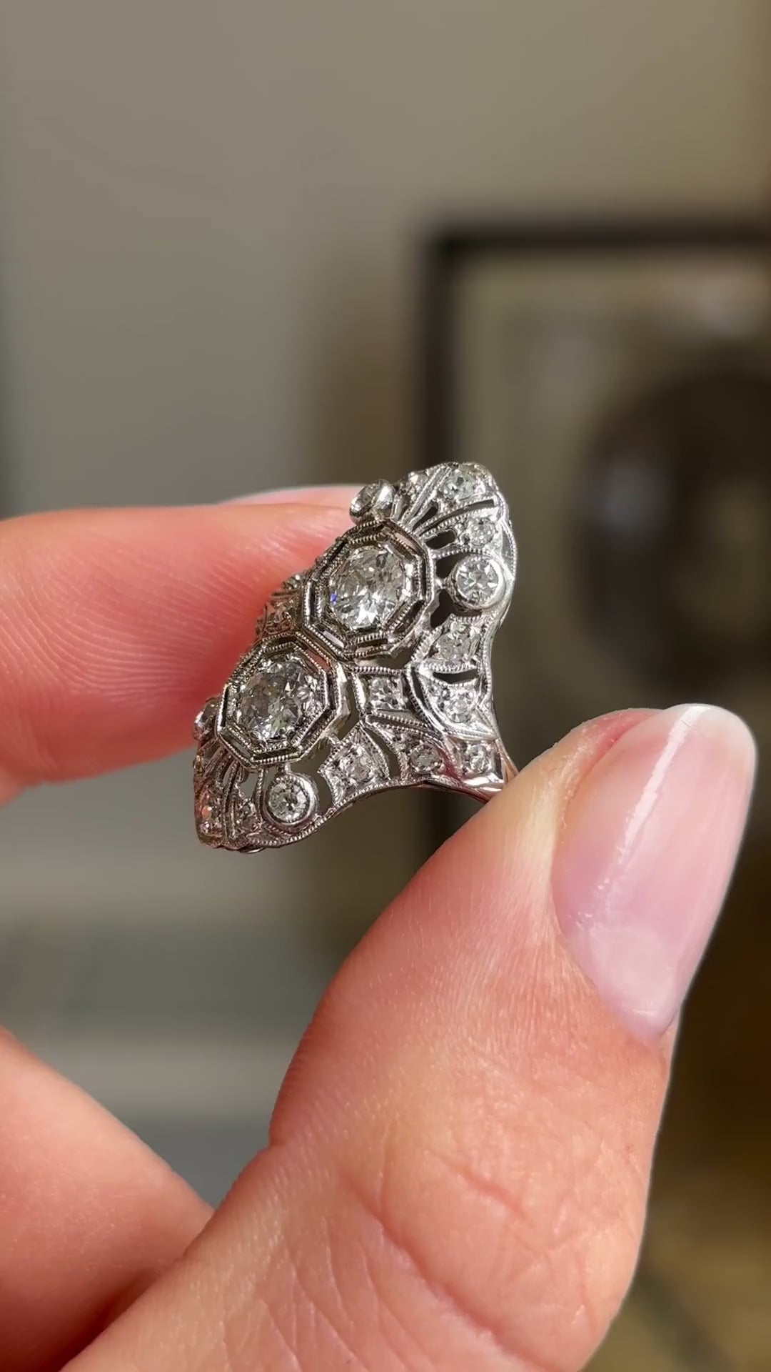 Detailed video of an antique Art Deco diamond cluster cocktail ring in platinum, held between fingers to showcase intricate filigree metalwork, old-cut diamonds and elegant 1920s craftsmanship against a warm interior setting.