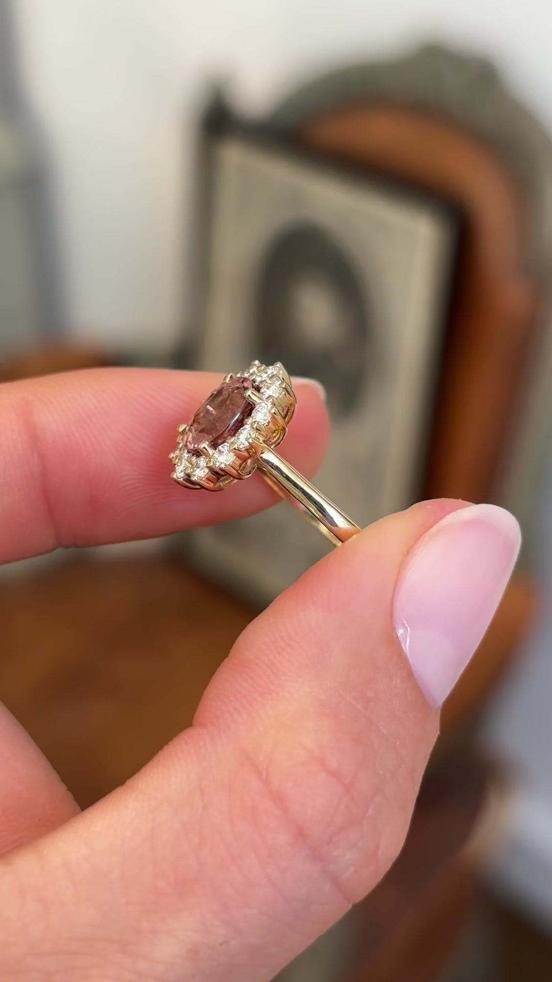 Antique-inspired rose-pink tourmaline and diamond cluster engagement ring featuring an oval rose-pink tourmaline encircled by brilliant-cut diamonds, held by hand for detailed viewing.