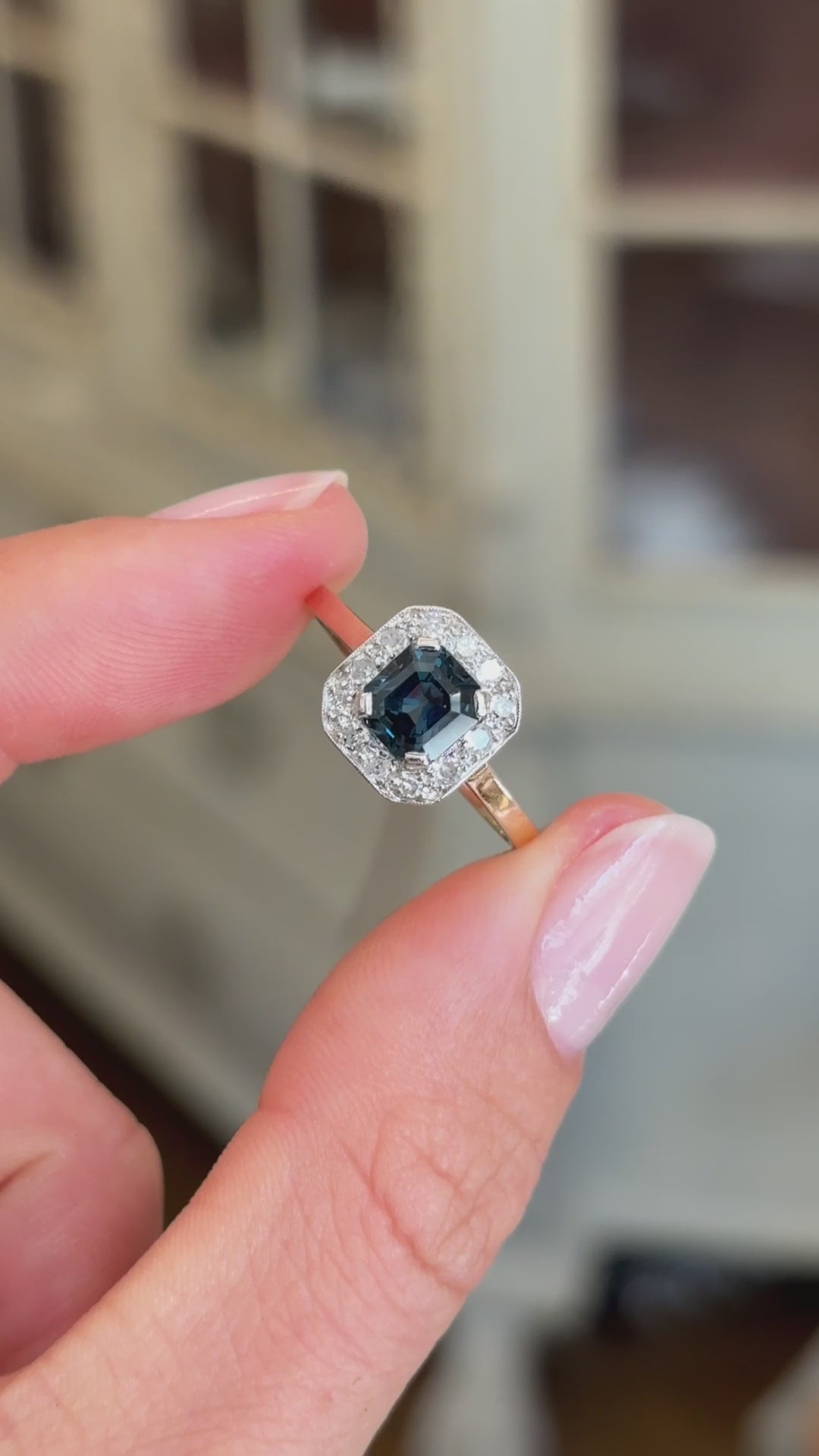Antique Art Deco sapphire and diamond cluster engagement ring featuring a blue sapphire centre stone surrounded by a geometric diamond cluster, held by hand for detailed viewing.