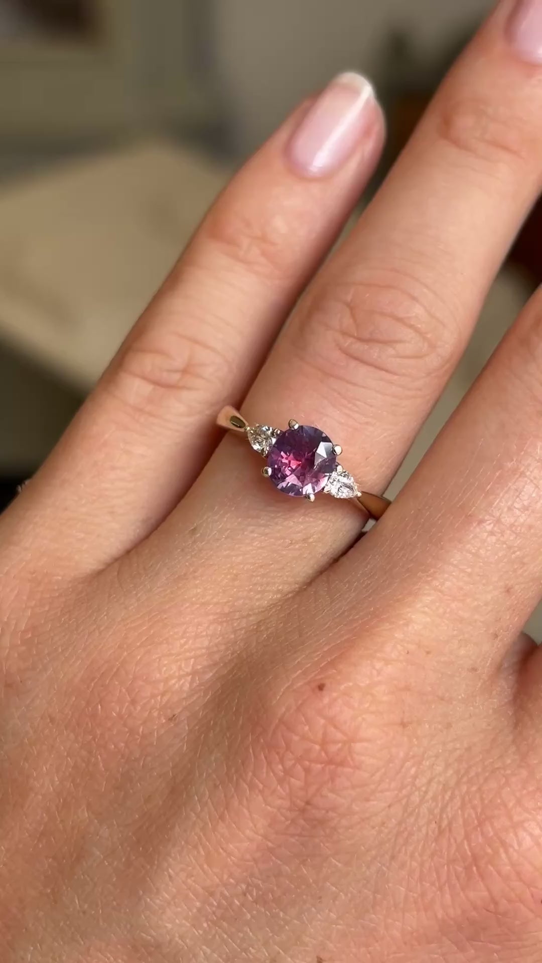 Pink-red sapphire and diamond three-stone engagement ring featuring a round pink-red sapphire centre stone with pear-cut diamond side stones, displayed on a hand.