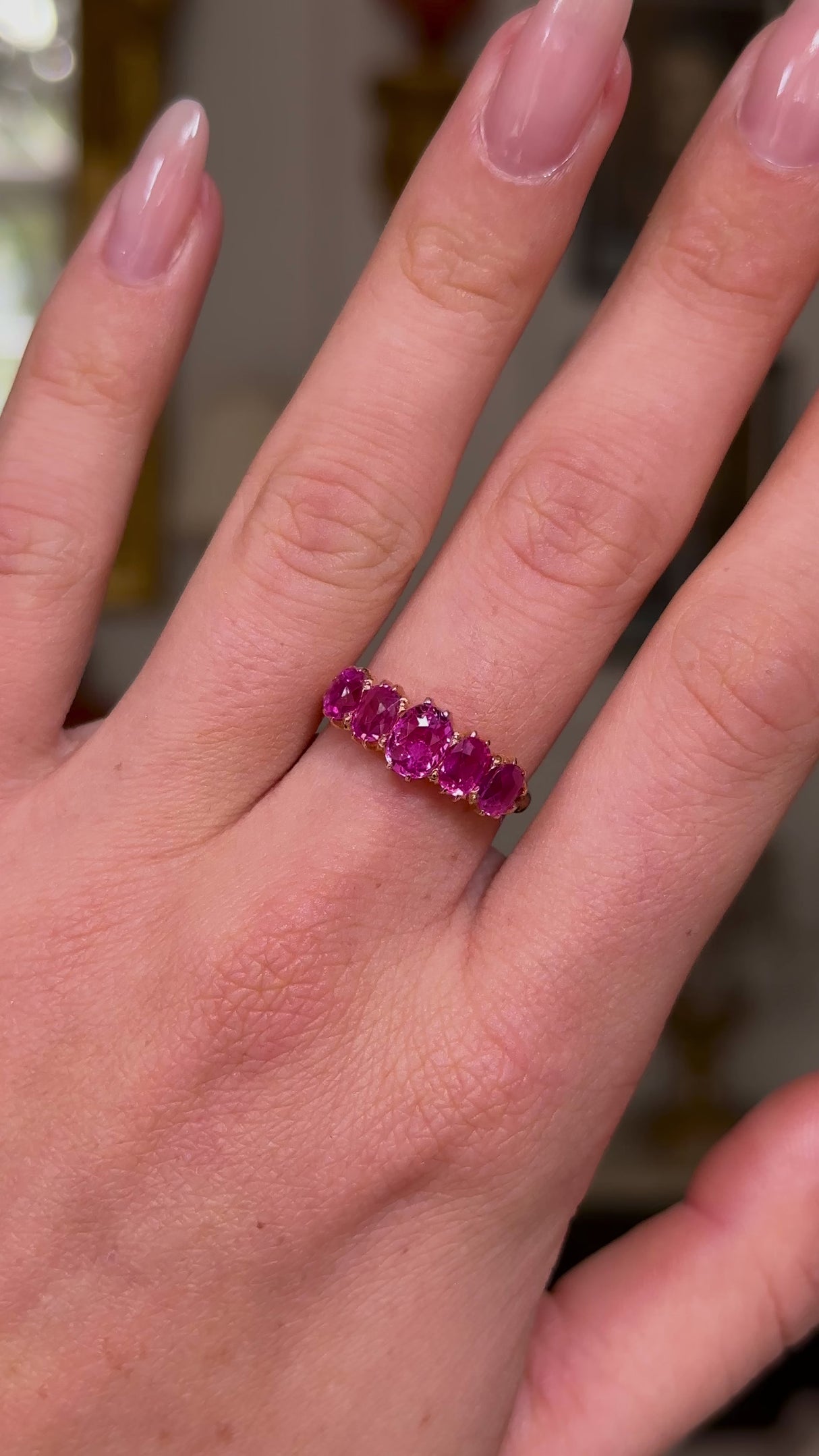 Five stone pink sapphire and yellow gold ring, worn on left hand middle finger, hand moving backwards from lens shot on antiquated bokeh background.