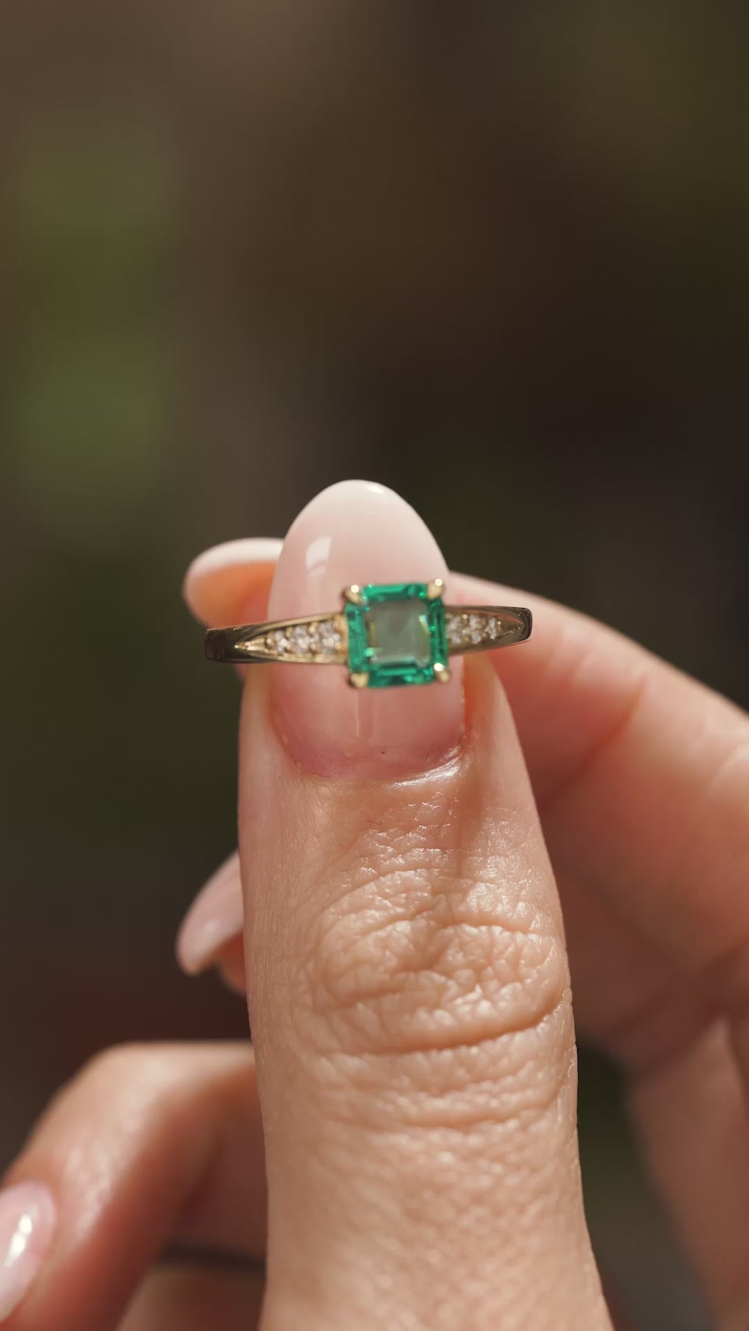 A video of a Vintage-inspired, Emerald and diamond flanked solitaire engagement ring in yellow gold held in between two fingers and slowly rotated to showcase different angles