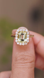 A close-up video of an Antique-inspired, Green beryl and diamond cluster engagement ring in yellow gold.  Held between two fingers and rotated slowly to show the basket setting and yellow gold band. 