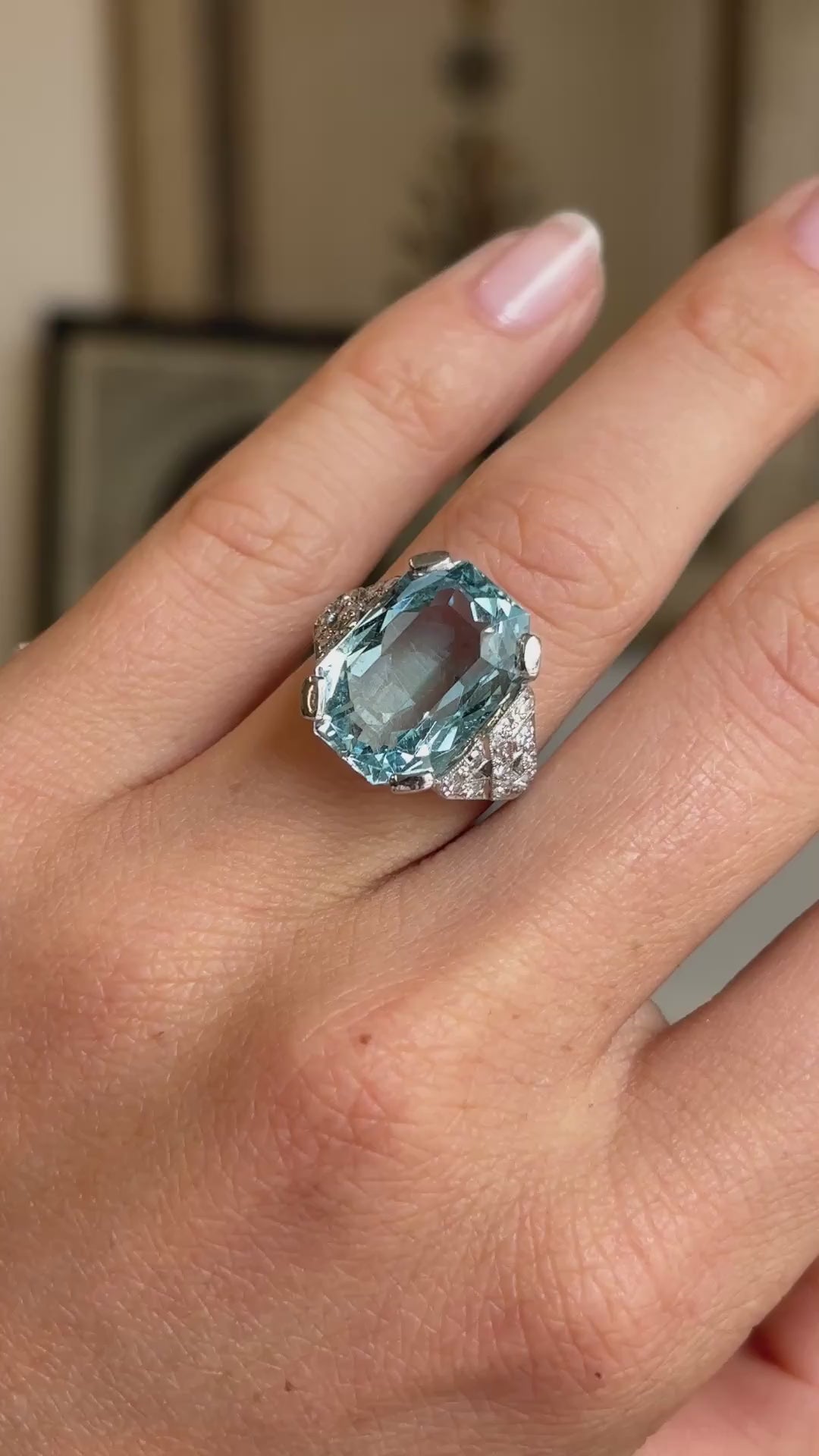 Video of an antique Art Deco aquamarine and diamond cocktail ring in platinum worn on hand, showcasing the vibrant blue aquamarine and sparkling diamond cluster against a warm vintage interior backdrop.