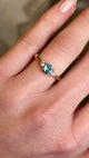 Round teal-green sapphire and diamond trilogy engagement ring worn on hand.
