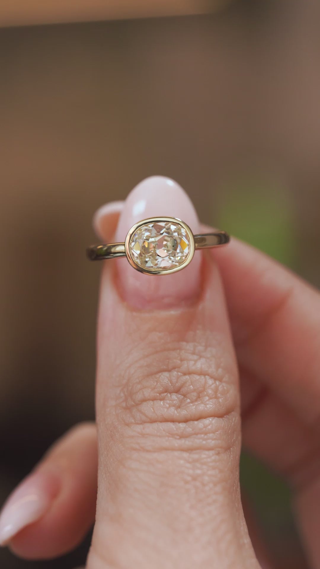 Video close-up of an antique-inspired diamond solitaire balanced on a fingertip, displaying the old-mine cut and polished gold surround.
