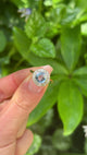 Vintage, cornflower blue sapphire & diamond cluster engagement ring held in fingers.