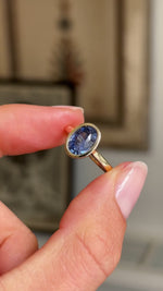 Detailed video of an antique-inspired oval-cut slate blue sapphire solitaire ring in yellow gold, held between fingers to showcase the smooth rub-over setting and beautifully saturated blue gemstone against a warm interior background.