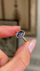 Detailed video of an antique-inspired oval-cut slate blue sapphire solitaire ring in yellow gold, held between fingers to showcase the smooth rub-over setting and beautifully saturated blue gemstone against a warm interior background.