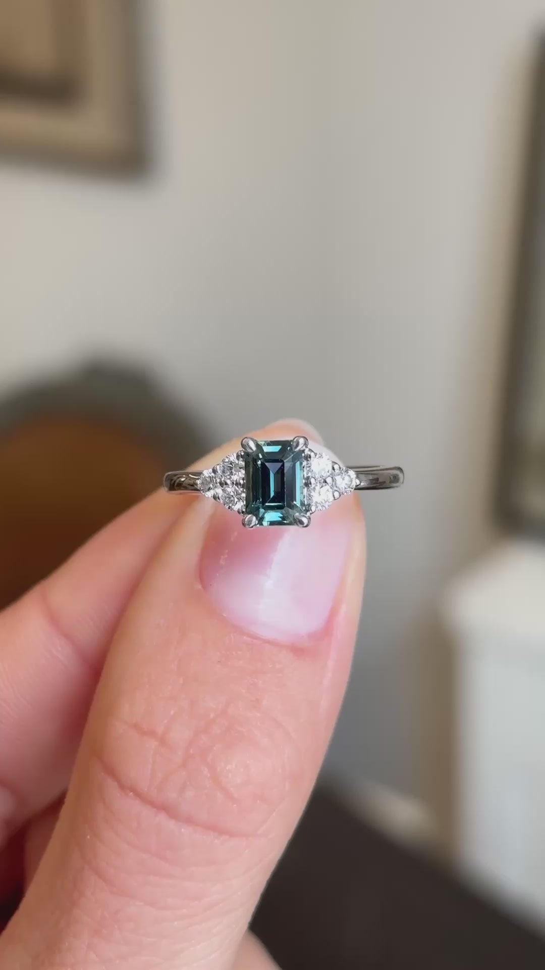 A video of an antique-inspired platinum engagement ring held between two fingers, showcasing a blue sapphire adorned with diamond trefoil clusters.