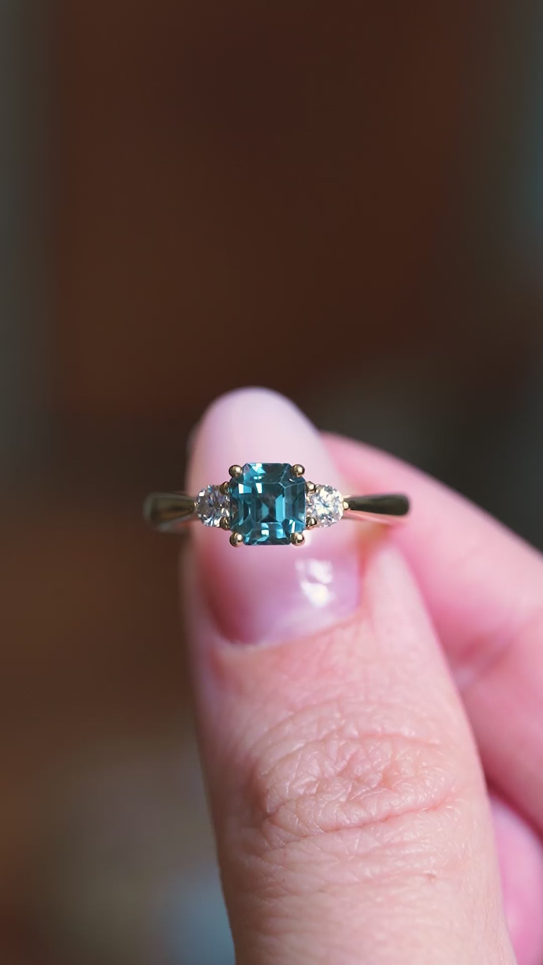 Teal sapphire and diamond three-stone engagement ring held between fingers showing ring details.